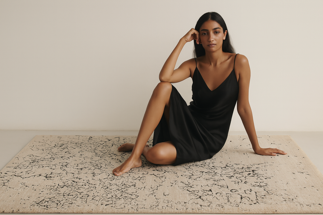 Woman in a black dress sitting on a patterned rug against a plain wall